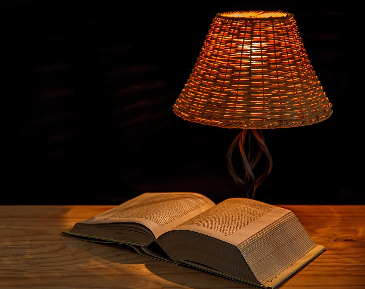 Lampe de chevet en rotin allumée sur table en bois avec livre ouvert – éclairage chaleureux pour ambiance de lecture cosy.