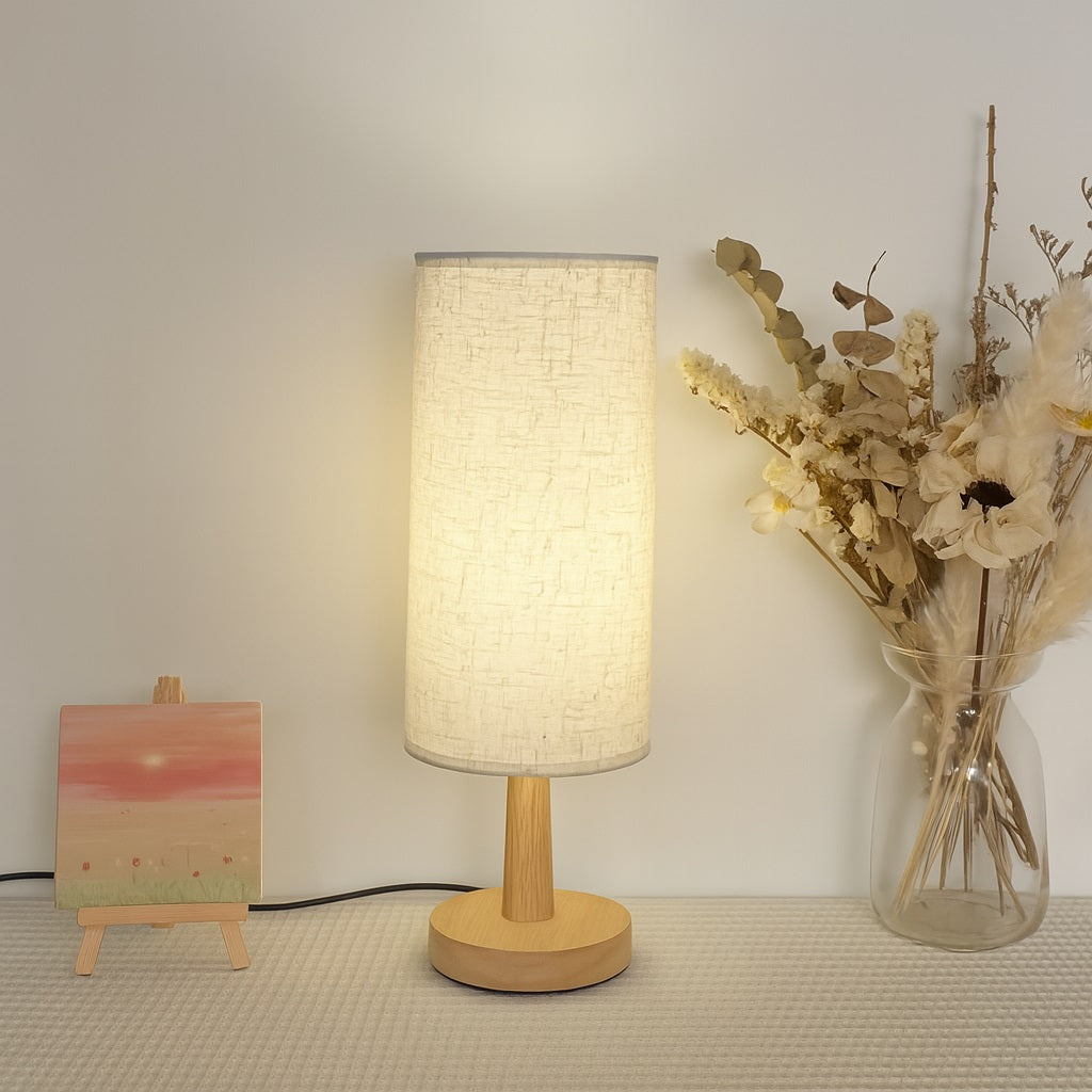 Abat-jour cylindrique blanc allumé sur lampe de chevet en bois, ambiance naturelle avec fleurs séchées en arrière-plan.