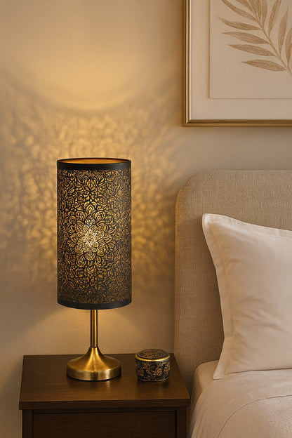 Abat-jour cylindrique en métal ajouré noir et doré, diffusant une lumière chaude et décorative sur table de chevet.