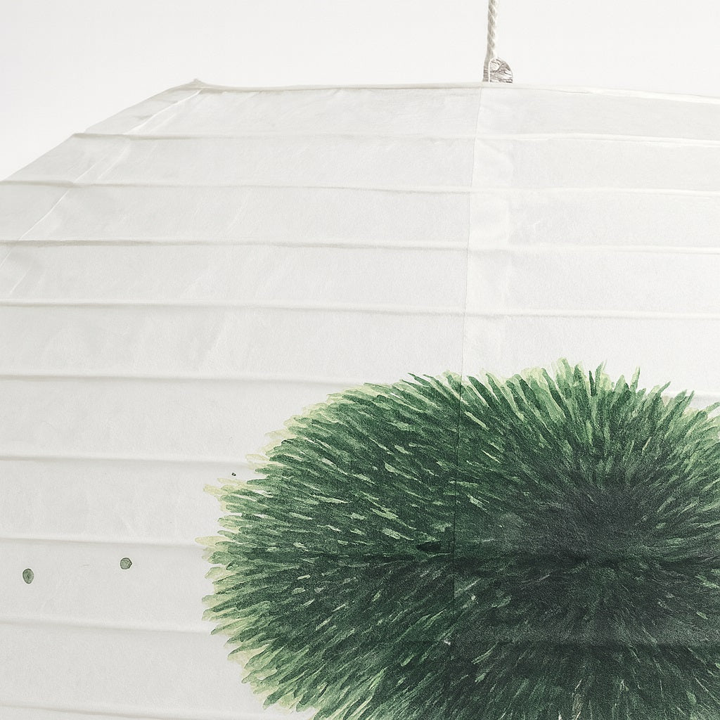 Détail d’un abat-jour en papier peint à la main, feuillage vert du bonsaï sur fond blanc texturé.